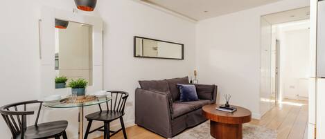 A modern living room with a glass dining table, two black chairs, a grey sofa with a blue pillow, a wooden coffee table, and a hanging pendant light.