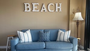A blue sofa with white and patterned cushions, a wall-mounted word "BEACH", a floor lamp, and a starfish decoration.