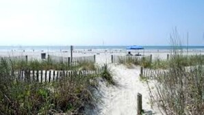 A sandy beach path leading to the ocean, with a fence and some vegetation in the foreground.