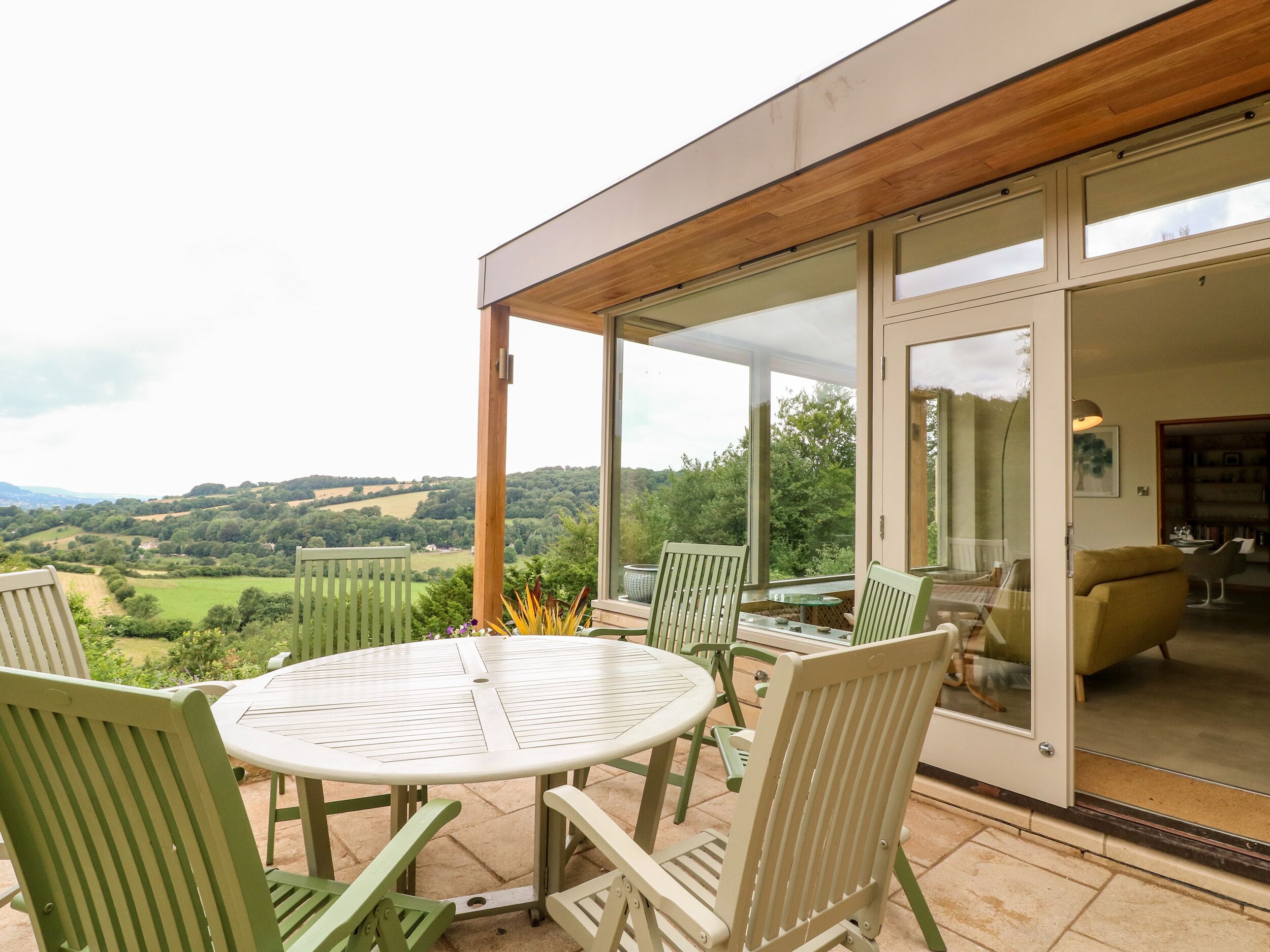 Eine Terrasse mit rundem Tisch und Stühlen, eine Glastür, die zu einem Innenbereich führt, und ein Blick auf die Landschaft.
