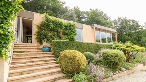 A modern house with a stone staircase leading to a glass door, surrounded by well-maintained hedges and a gravel path. - Under Catswood (Stroud)