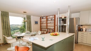 A modern kitchen with a central island, bar stools, a dining area with a view, and a built-in bookshelf. - Under Catswood (Stroud)