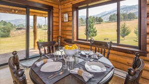 Un comedor de madera con una mesa preparada para cuatro personas, grandes ventanales que dan a un campo cubierto de hierba y montañas.