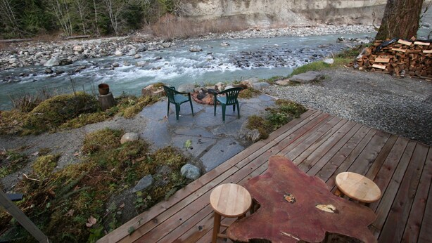 Una terraza de madera con dos sillas y una mesa, con vista a un río con rocas y una pila de leña.