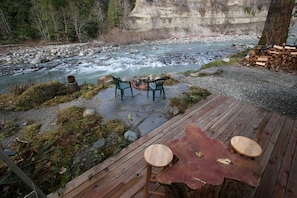 Une terrasse en bois avec deux chaises et une table, donnant sur une rivière bordée de rochers et sur laquelle se trouve une pile de bois de chauffage.