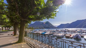 A marina with boats docked, a tree-lined walkway, and mountains in the background. - Lugano Terrazzo (Lugano)