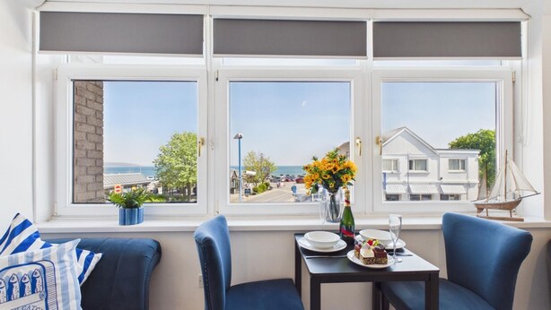A dining area with a table set for two, a window with a view of the sea and buildings, and a potted plant on the windowsill. - Ocean View - 1 Bedroom - Saundersfoot (Saundersfoot)