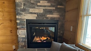 A cozy living room with a stone fireplace, a gray sofa, and a window with blinds. - #1 Cabin~Walk to WISP! Lake access & dock! HOT Tub~Game room~Fire Pit & DOGS OK! (McHenry)