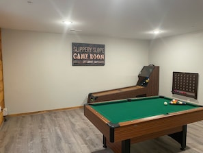 A pool table with a green felt surface, a pool cue, and several balls. There is a sign on the wall that reads "Slippery Slope Game Room." The room has wooden flooring and a white wall.