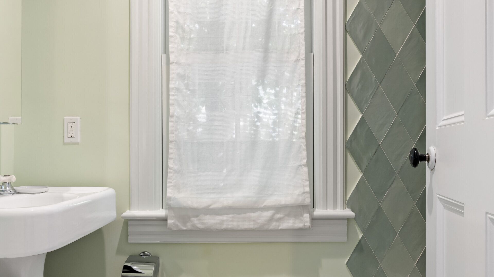 A bathroom with a white sink, a window with a white curtain, and a door with green diamond-patterned tiles.