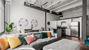 A modern living room with a grey sectional sofa, a grey ottoman, and a dining area with white cabinets and a stainless steel refrigerator.