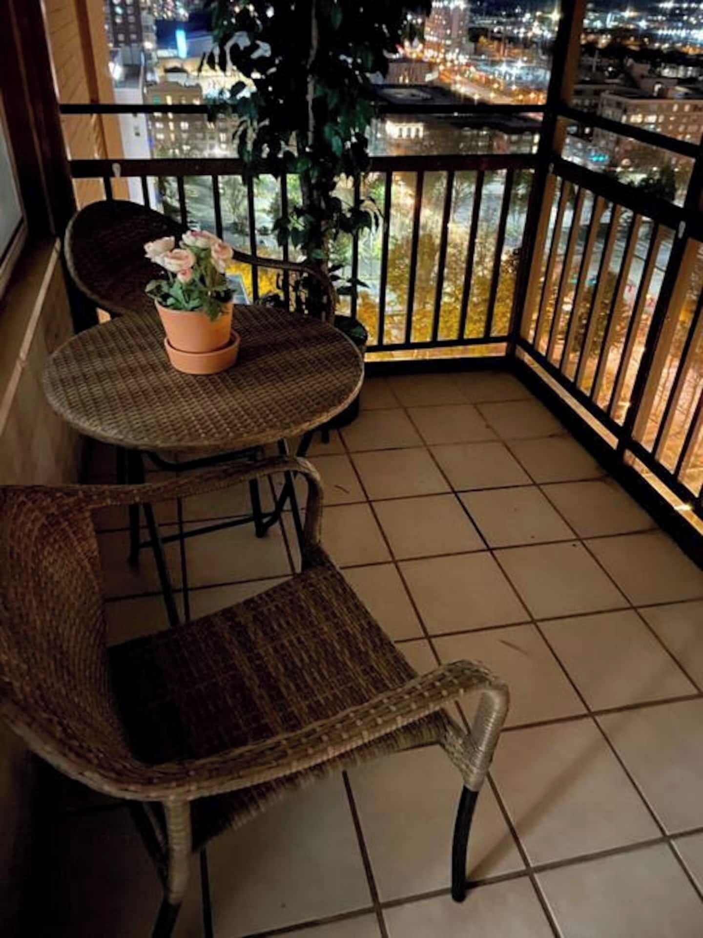 A balcony with a round table and two wicker chairs, a potted plant with flowers, and a city view at night.