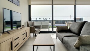 A modern living room with a flat-screen TV, a sofa, a coffee table, and a view of a pool and palm trees through large windows. - Modern villa with private pool (Mar de Cristal)