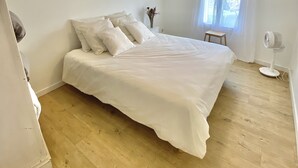 Ein Schlafzimmer mit einem großen Bett, einem kleinen Hocker, einem Standventilator und einem Fenster mit Vorhängen.