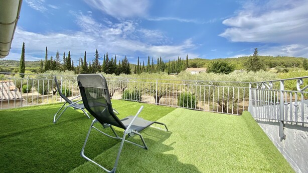 Eine Terrasse mit Liegestuhl, einem weißen Metallzaun und Blick auf eine ländliche Landschaft mit Bäumen und einem Gebäude in der Ferne.
