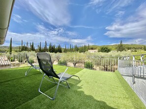 A patio with a lawn chair, a white metal fence, and a view of a countryside landscape with trees and a building in the distance.
