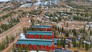 A multi-story red building with a green roof, surrounded by a forested area with snow patches, and a mountain in the background. - JUST FINISHED RENOVATIONS 2023!!! A lavishly styled ski-in/out winter escape! (Brian Head)