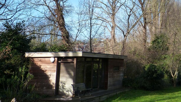 A small wooden cabin with a porch, surrounded by trees and grass. - Highgate Studio (London)