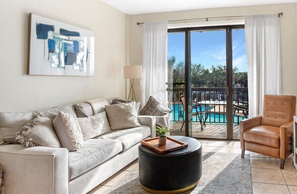 A living room with a sofa, a round ottoman, a chair, and a pool view.