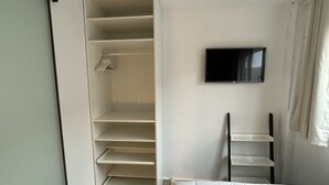 A hotel room with a bed, a TV mounted on the wall, a closet with shelves, and a window with curtains.