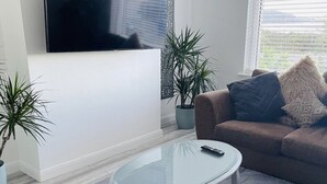 A living room with a flat-screen TV mounted on the wall, a round glass coffee table, a brown sofa with cushions, and two potted plants.