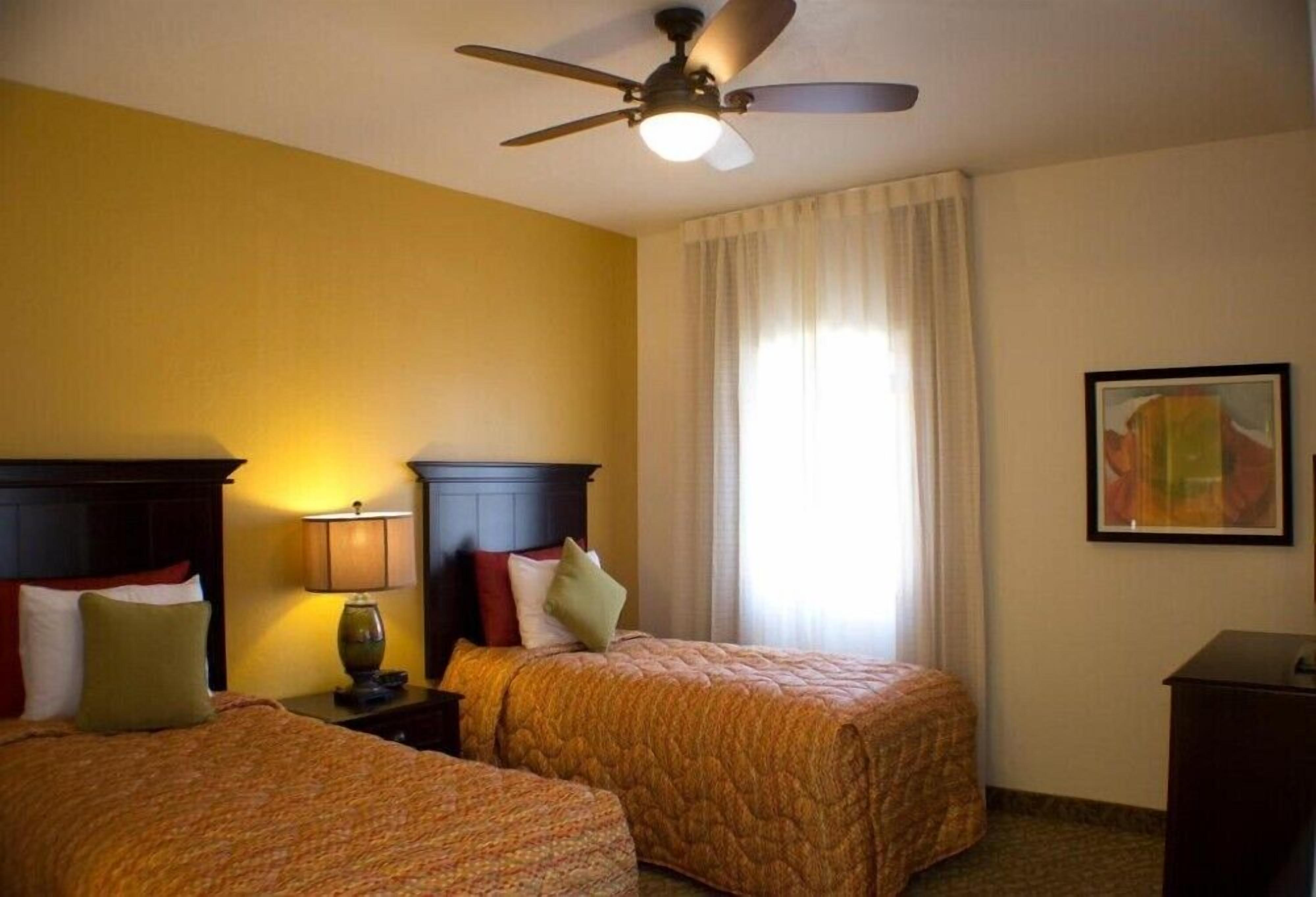 Habitación de hotel con dos camas, un ventilador de techo, una mesita de noche con lámpara y un cuadro en la pared.