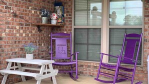 A patio with a white picnic table and two purple rocking chairs, a brick wall, and a window with blinds. - Peaceful 3BR/2BA Hidden Lake Home Near ACOM w/WiFi (Dothan)