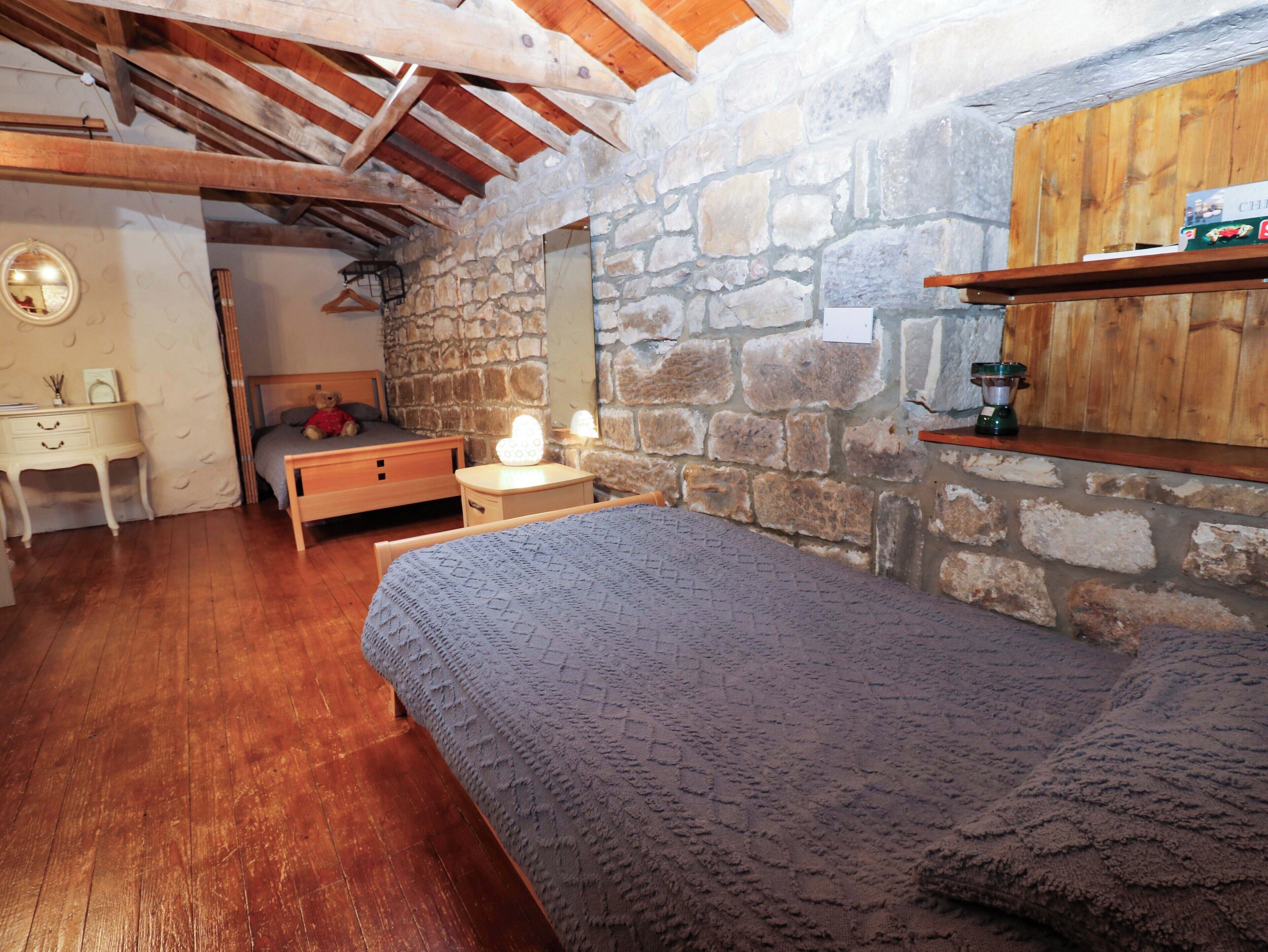 A cozy bedroom with a stone wall, wooden beams, a bed, a desk, and a nightstand.