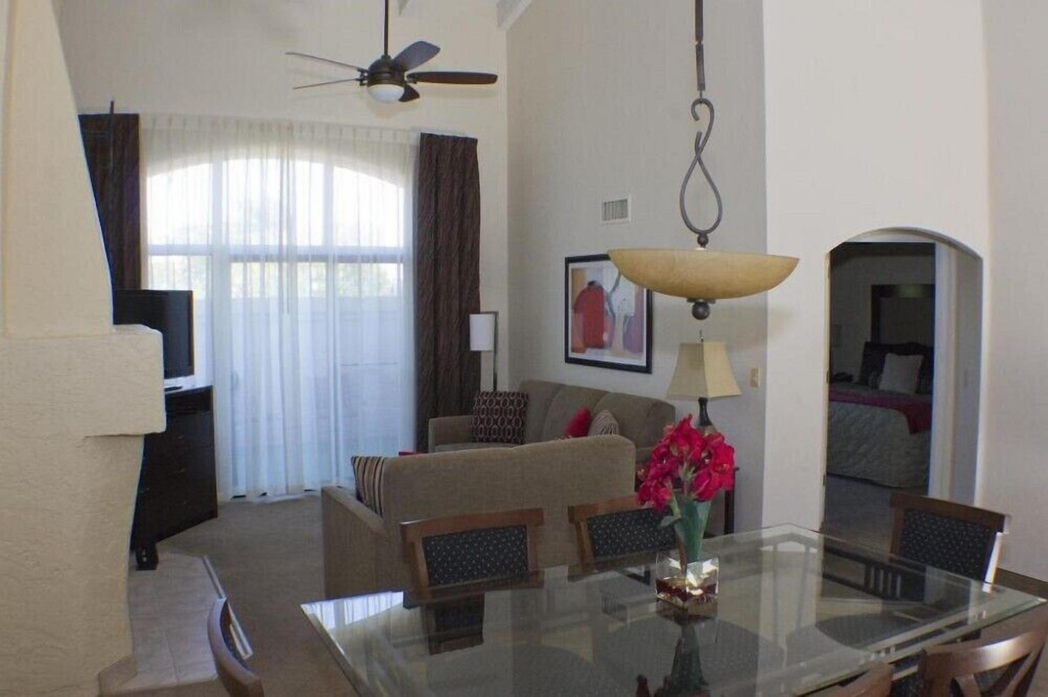 A hotel room with a dining table, chairs, a sofa, a ceiling fan, and a hanging lamp.