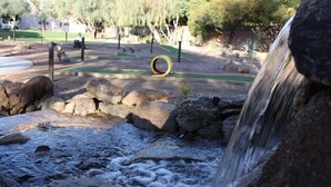 Um parque com vegetação exuberante, uma cascata, pedras e um parquinho circular.