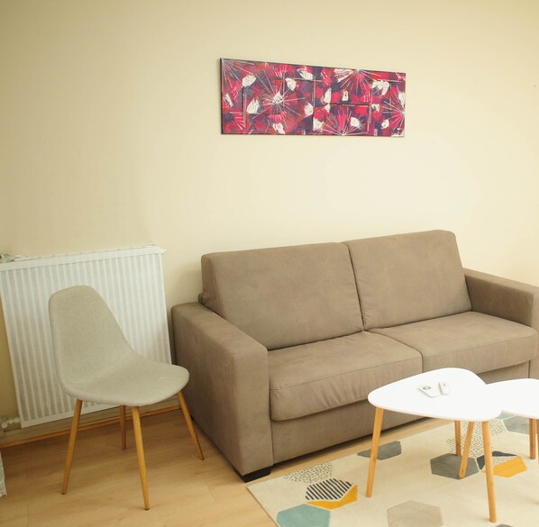 A living room with a beige sofa, a grey chair, a white coffee table, and a radiator. - Spacious, modern T3, near metro and RER station (Créteil)