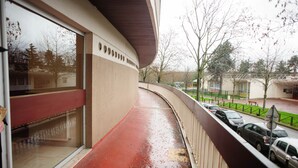 En rød balkon med glasdør, buet gelænder og udsigt til parkerede biler og træer.