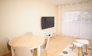 A modern living room with a round wooden table, grey upholstered chairs, a flat-screen TV mounted on a white cabinet, and a large window with sheer curtains.