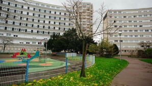 En legeplads med rutchebane og gynger, omgivet af et hegn, foran en høj boligbygning.