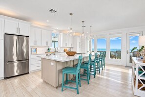 A modern kitchen with a stainless steel refrigerator, white cabinetry, a large island with bar stools, pendant lighting, and a view of the ocean through large windows. - Beachfront Retreat with SwimSpa | Putt-Putt | Fire Pit | Sleeps 16 (Bolivar Peninsula)