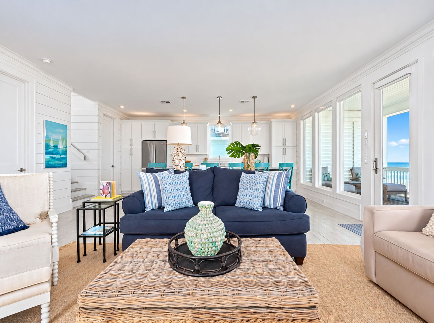 A modern living room with a dark blue sofa, a wicker coffee table, and a view of the ocean through large windows.