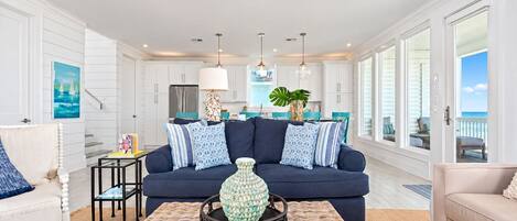 A modern living room with a dark blue sofa, a wicker coffee table, and a view of the ocean through large windows.