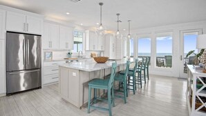 Une cuisine moderne avec des meubles blancs, un réfrigérateur en acier inoxydable et un grand îlot central agrémenté de chaises de couleur turquoise.