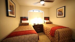 A hotel room with two single beds, a nightstand with a lamp, and a ceiling fan.