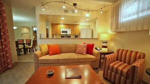 A living room with a sofa, armchair, coffee table, and a kitchen in the background.