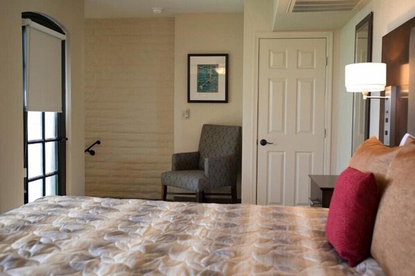Habitación de hotel con cama, una silla, una puerta, un cuadro enmarcado en la pared y una lámpara.