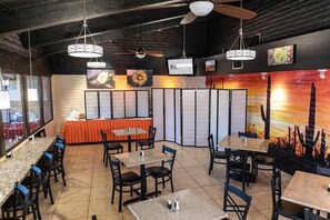 Interior de restaurante con varias mesas y sillas, ventiladores de techo y televisores en la pared.