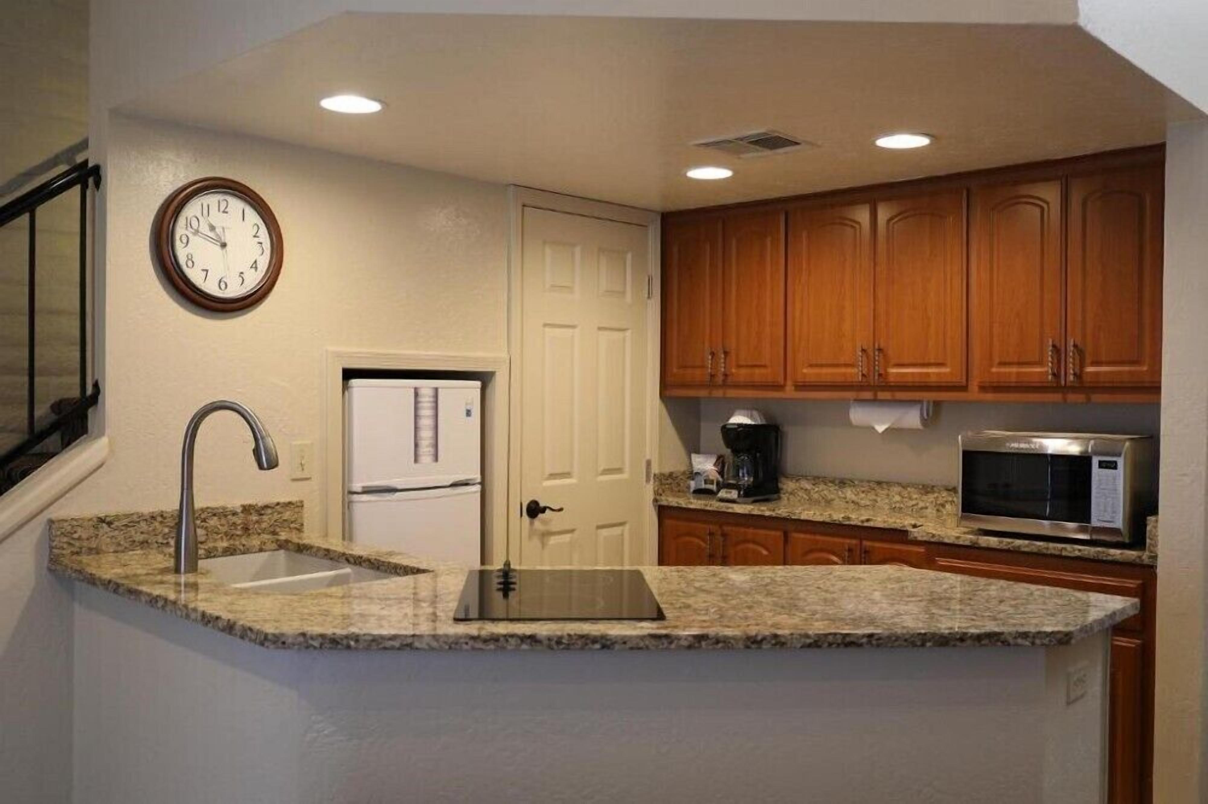 A kitchen with granite countertops, wooden cabinets, a stainless steel microwave, a coffee maker, and a clock on the wall.