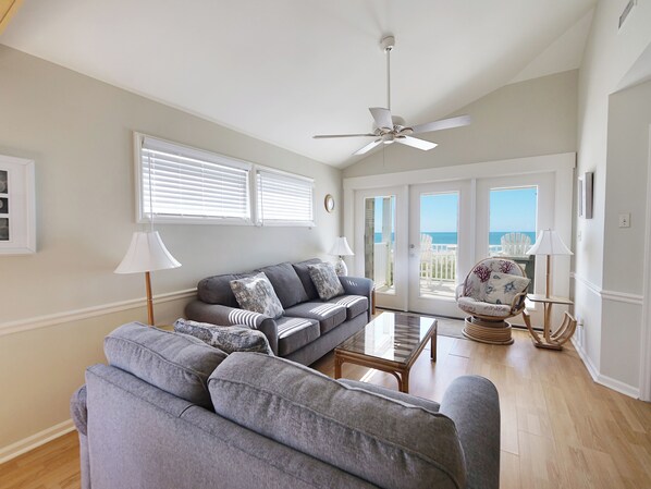A spacious living room with a grey sofa, a coffee table, and a ceiling fan. There are two windows with blinds, a door leading to a balcony, and a view of the ocean.