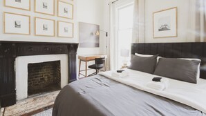 A hotel room with a fireplace, a bed with grey bedding, a desk with a chair, and framed pictures on the wall.