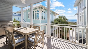 A covered outdoor deck with a table and chairs, a ceiling fan, and a view of a light blue house with a balcony. - Gulf Front Views | "All Decked Out" Luxurious WaterColor Home | Steps to the Beach Club (Santa Rosa Beach)