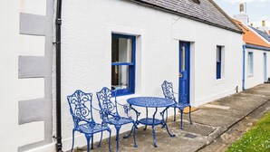 Un bâtiment blanc avec des portes et des fenêtres bleues, un toit en tuiles et une petite table d’extérieur entourée de chaises en métal bleues.