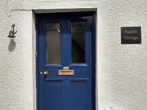 A blue door with a window, a brass knocker, and a sign reading 'Needle Cottage.'
