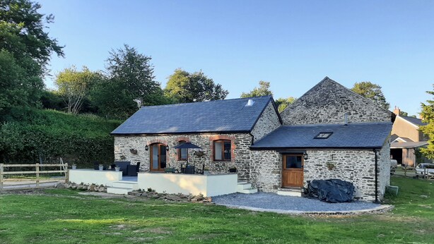 A stone house with a sloped roof, a front porch with outdoor seating, and a small attached shed. - Slade Cottage - UK35027 (Brayford)
