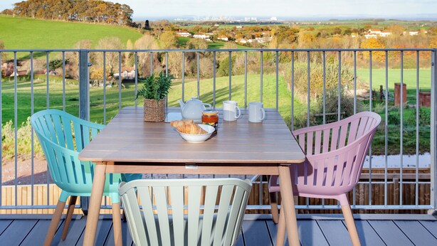 Une table en bois, entourée de chaises, sur un balcon donnant sur un paysage rural.